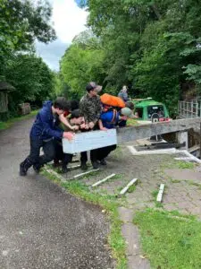 Group of youths leaning on a white wooden railing beside a canal, preparing to board a green boat in a leafy outdoor setting.