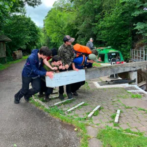 Group of people in outdoor gear push a long blue-painted barrier along a paved path by trees.