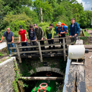 Group of six people outdoors on a wooden footbridge over a canal lock, wearing backpacks and jackets.