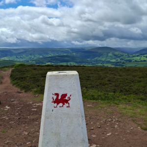 White milestone pillar in the foreground with a red Welsh dragon, set against rolling green hills and a cloudy sky.