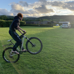 Person riding a bicycle across a green field with rural buildings and hills in the background under a cloudy sky.
