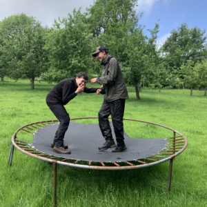 Two people on a trampoline outdoors in a grassy park, smiling and leaning toward each other amid green trees on a cloudy day.