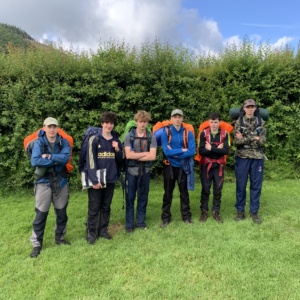 Group of young hikers standing side by side with backpacks on a grassy field, green hedge behind them and blue sky above.
