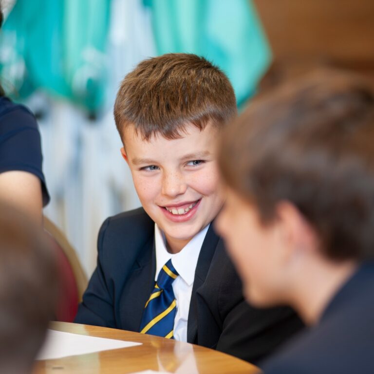 Smiling boy in a navy blazer and striped blue-yellow tie sits at a table, talking with a classmate.