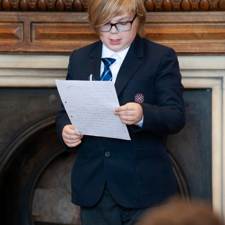 Boy in a school uniform reads a handwritten letter in front of a fireplace, wearing glasses and a navy blazer.