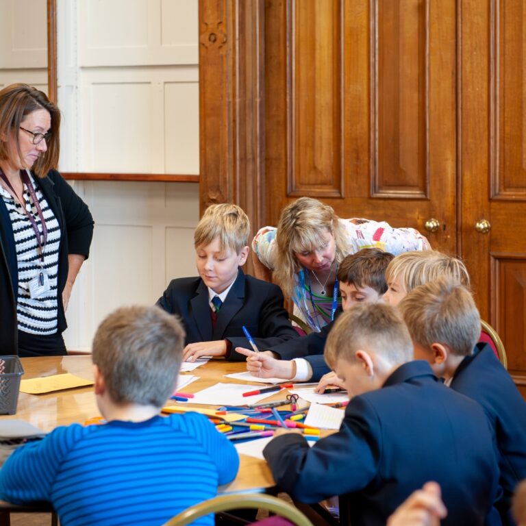 Teacher and an adult supervise several schoolchildren as they draw and write at a table.