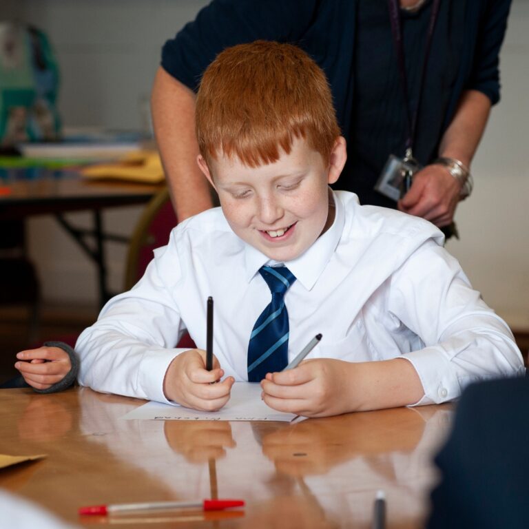 Red-haired boy in a white shirt and striped tie writes on a paper at a desk, with a teacher standing behind supervising.
