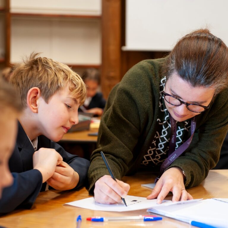 Teacher leans over a table to help a student with writing on a worksheet in a classroom setting.