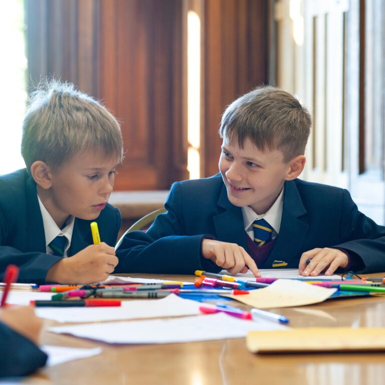 Two schoolboys in navy blazers work together at a table scattered with colorful markers and papers, smiling as they draw and discuss a project.