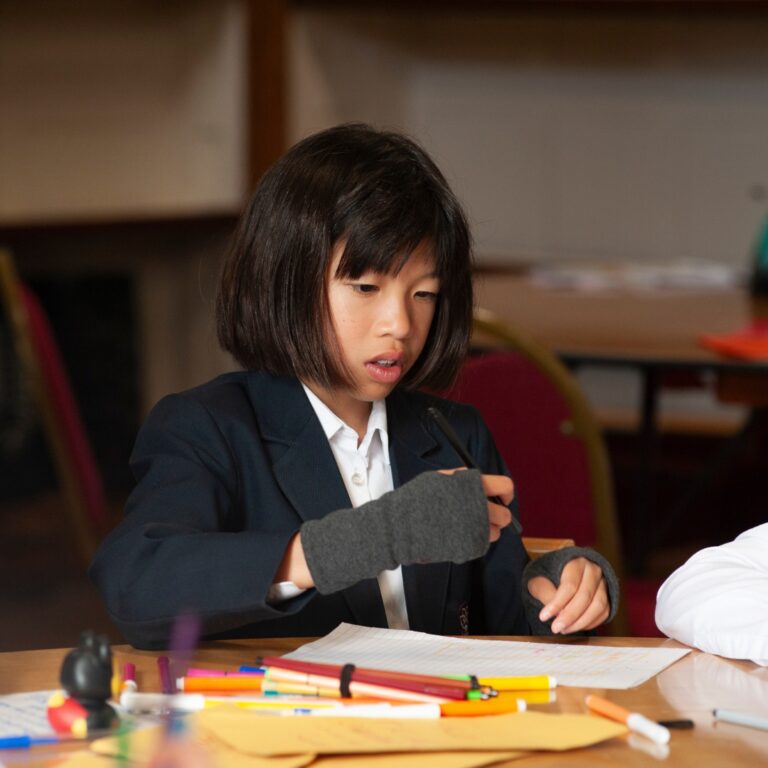 Girl in a navy blazer sits at a table drawing with colored pencils and paper in front of her.