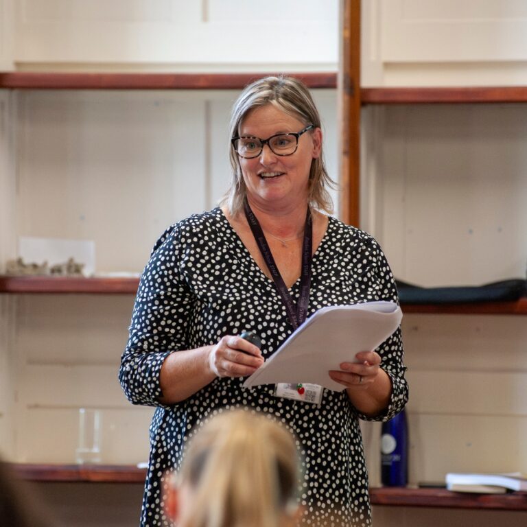Woman with glasses giving a presentation, holding papers and a remote in a polka-dot blouse.