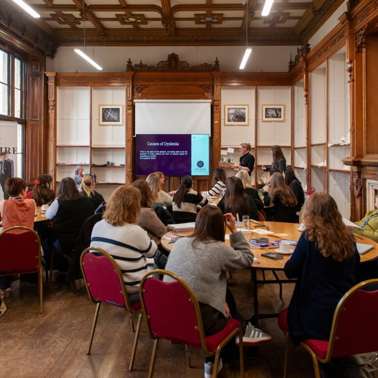 Group of adults seated around round tables in a wood-paneled lecture room, listening to a presentation on a screen reading 'Causes of Dyslexia'.