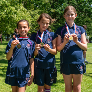 Three girls in navy school uniforms wear gold medals on red-white-blue ribbons, posing outdoors on green grass.