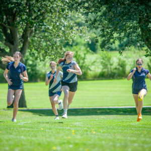 Group of young girls sprinting on a sunny grassy field during a race, wearing blue athletic uniforms.