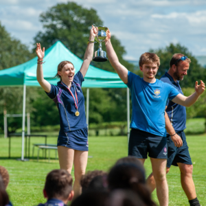Athletic team celebrates victory outdoors, raising a silver trophy as others cheer on a sunny day.