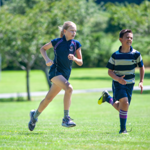 Two children running on a sunny grassy field in athletic uniforms, mid-stride in a race setting.