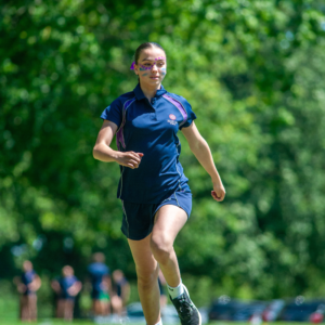 Athlete in navy uniform mid-jump outdoors with painted face and a green blurred background.