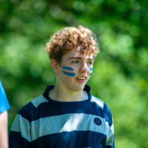 Young man with blue face paint stripes on cheeks, outdoors with green foliage in background, wearing a striped navy and light blue shirt.
