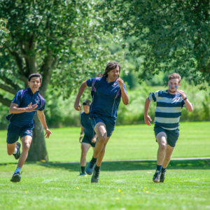Group of runners sprinting on a grassy field with trees in the background on a sunny day.