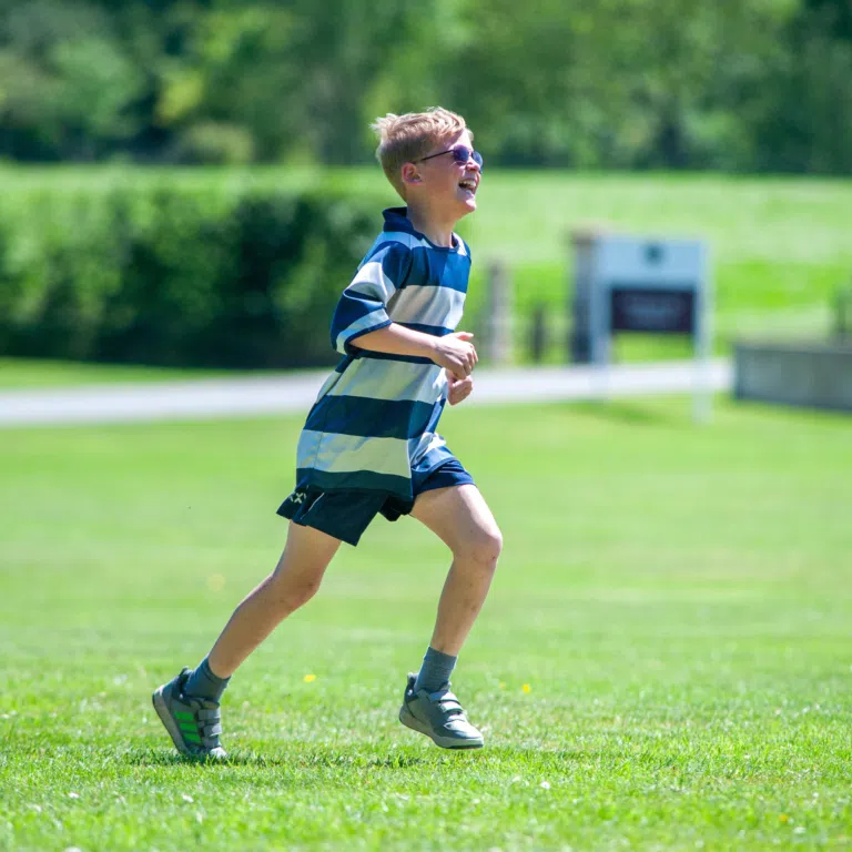 A child running on a sunny grassy field, wearing a blue and white striped shirt and sunglasses, smiling as they move.