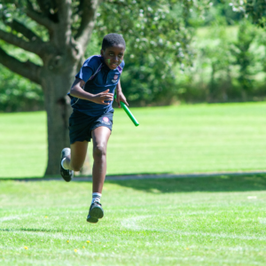 Child in a blue school uniform runs with a green baton across a sunny park lawn.