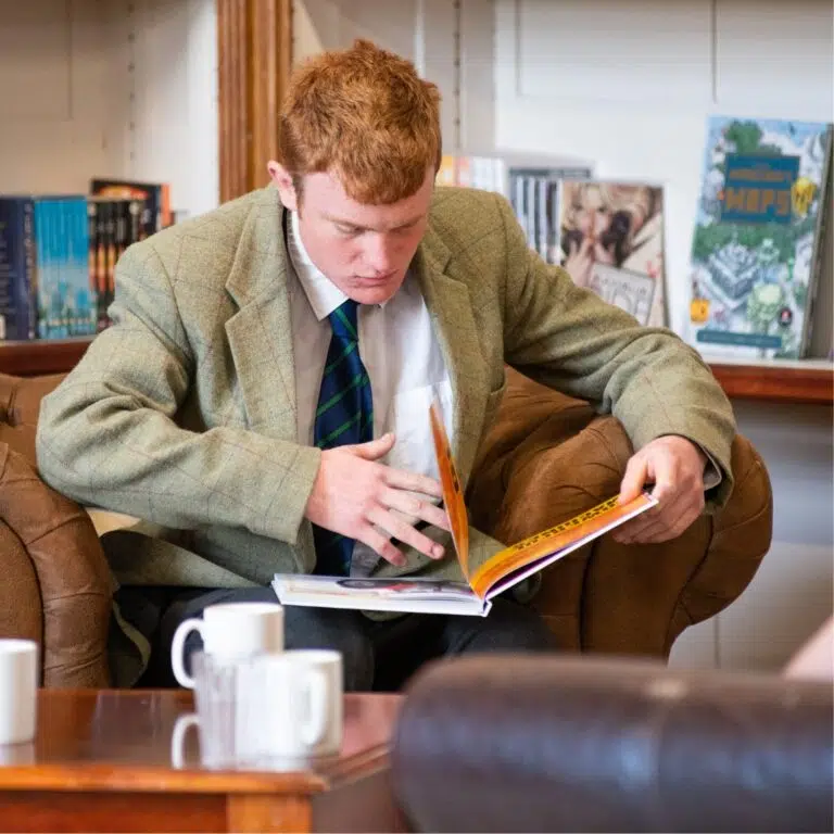 Man in a tweed blazer sits on a brown sofa reading a magazine in a library or(bookstore) lounge, concentrating on the pages.