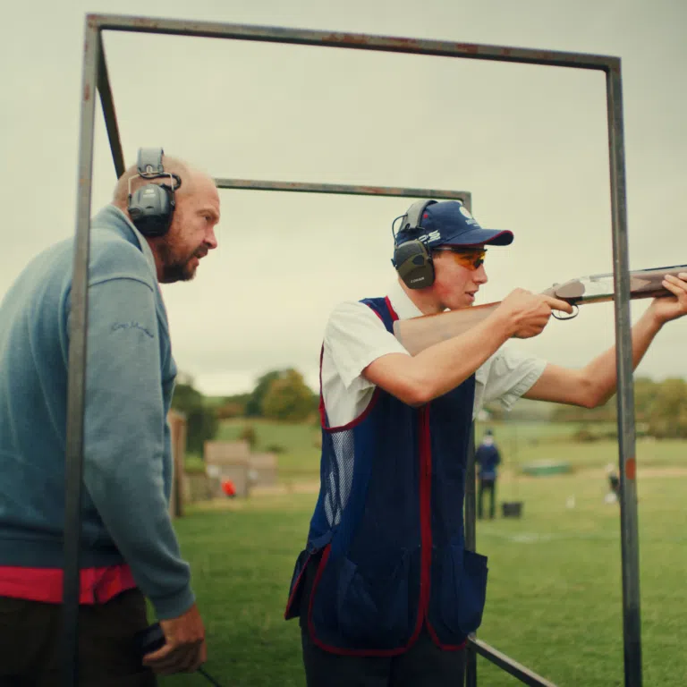 Two men wearing ear protection aim a rifle through a metal frame at a shooting range outdoors on a sunny day.