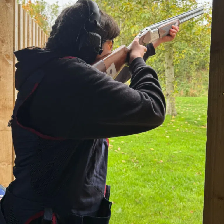 Person in a dark hoodie wearing protective earmuffs, aiming a shotgun outdoors beside wooden posts in a grassy area.