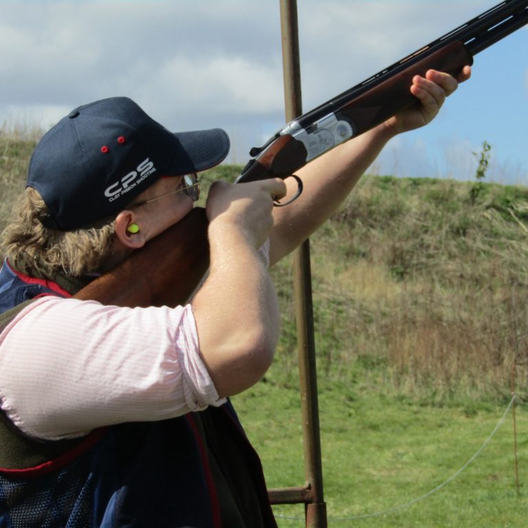 Person in a blue cap and ear protection aiming a rifle outdoors in a grassy field at arm's length from the camera.