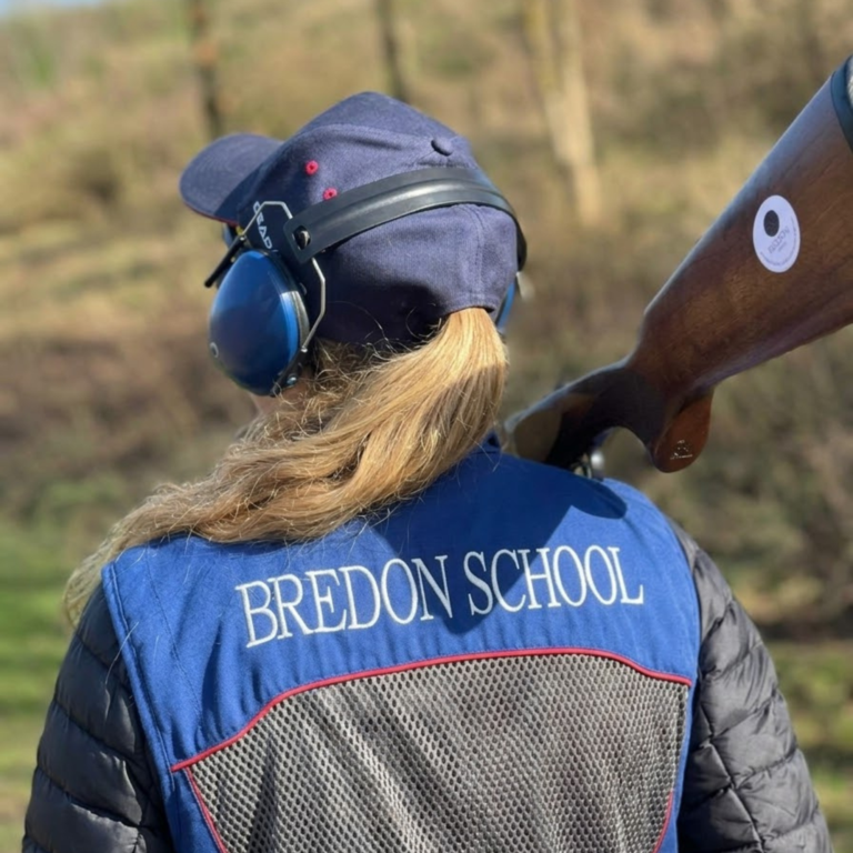 Rider in a blue vest with white letters on back, wearing a helmet and ear protection, seated on a brown horse outdoors.