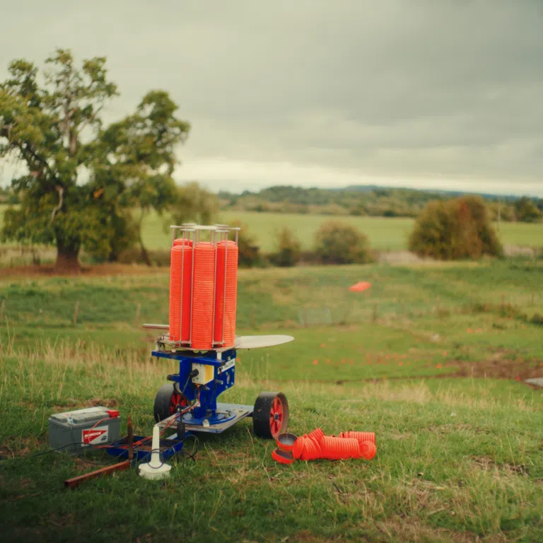 Orange model rocket launch tubes on a blue platform in a grassy field, with trees and hills in the blurred background.