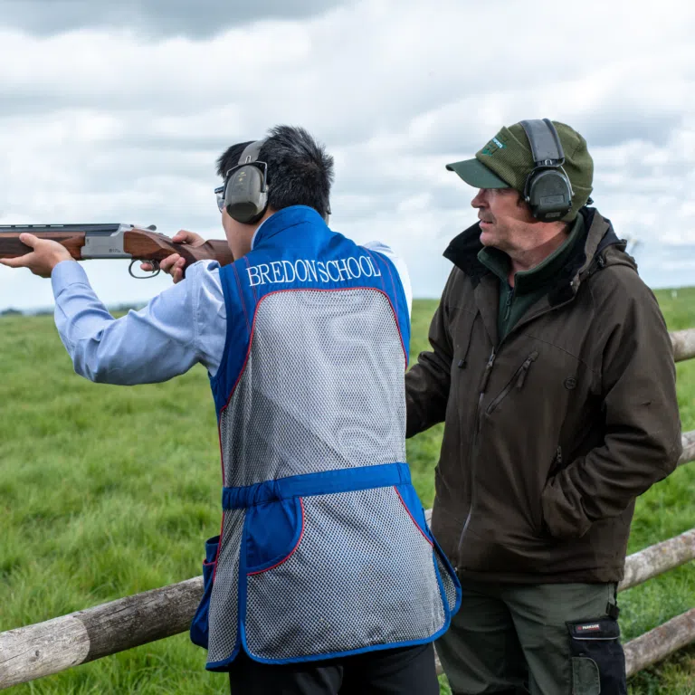 Two men outdoors beside a wooden fence; one in a blue shooting vest aiming a shotgun while the other watches, wearing a green cap and earmuffs.