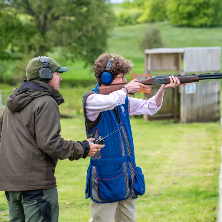 Two people at an outdoor shooting range; one guides the other who is aiming a rifle while wearing hearing protection and safety gear.