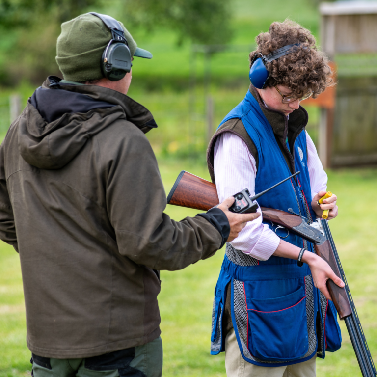 Two people outdoors, one man in a brown jacket facing away and another person in a blue vest and earmuffs holding a rifle and looking down at it.