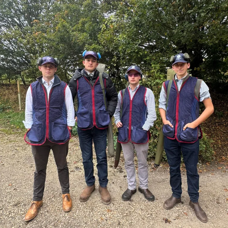 Five men wearing blue outdoor vests and sturdy boots stand together on a gravel path with trees in the background.