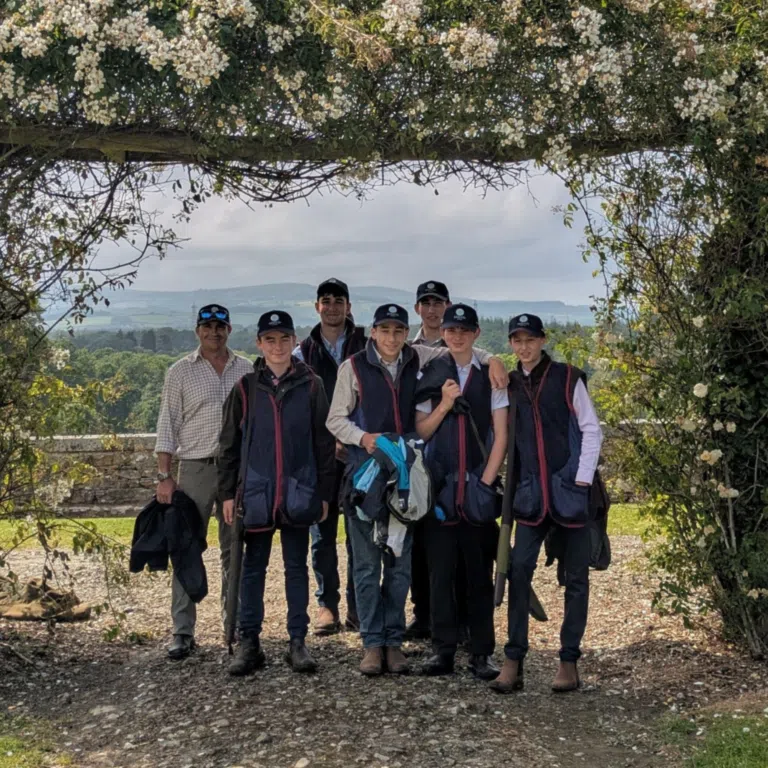 Group of young men in dark jackets and caps posing outdoors under a flowering vine arch.