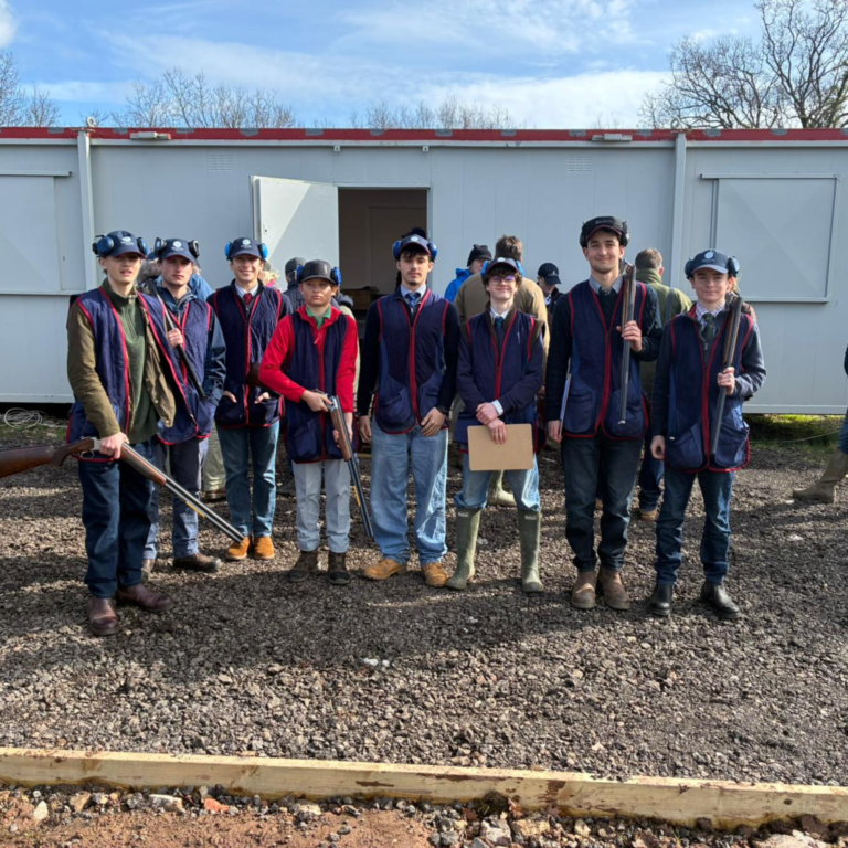Group of teens in blue vests and safety gear posing with rifles outdoors in a gravel lot near a light building.