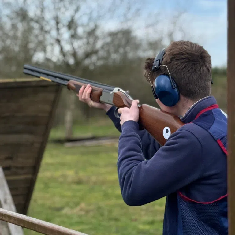 Person wearing ear protection aiming a shotgun outdoors, with the gun resting on their shoulder at a firing range or yard.
