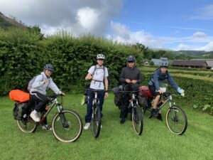Group of four kids with helmets posing with bicycles on a grassy field, hedges and hills in the background under a cloudy sky and blue sky.