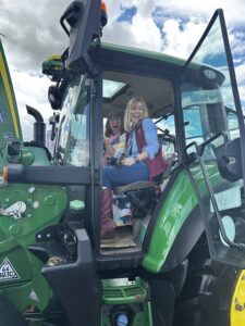 Two women sit in a green tractor cab, smiling and excited with the door open.