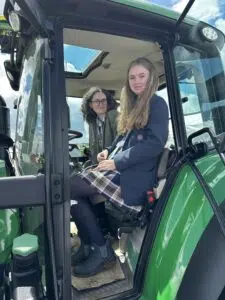 Two young women sit in the cab of a green tractor, smiling at the camera with a blue sky seen through the sunroof&hellip;]