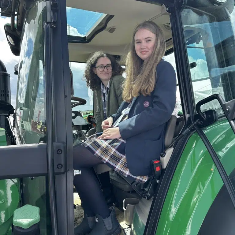 Two young women sit in the cab of a green tractor, smiling at the camera with a blue sky seen through the sunroof…]
