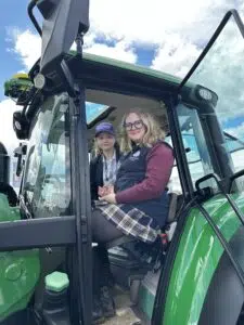 Two people sit in the cabin of a green tractor, smiling at the camera with the door open and blue sky visible outside.