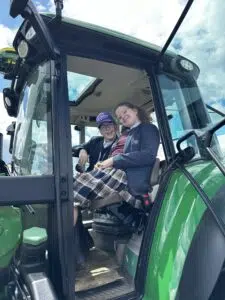 Two young girls in school uniforms sit in the driver&rsquo;s seat of a green tractor, smiling at the camera with the door open.