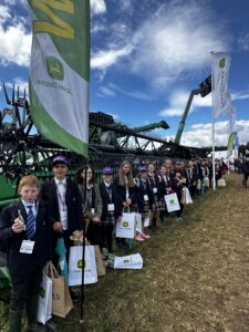 Line of students in school uniforms posing with John Deere machinery at an outdoor field event, each holding branded bags.