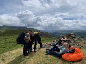 Group of hikers taking a break on a grassy hillside with large backpacks and a bright orange tarp on the ground. Clouds loom overhead over rolling hills.
