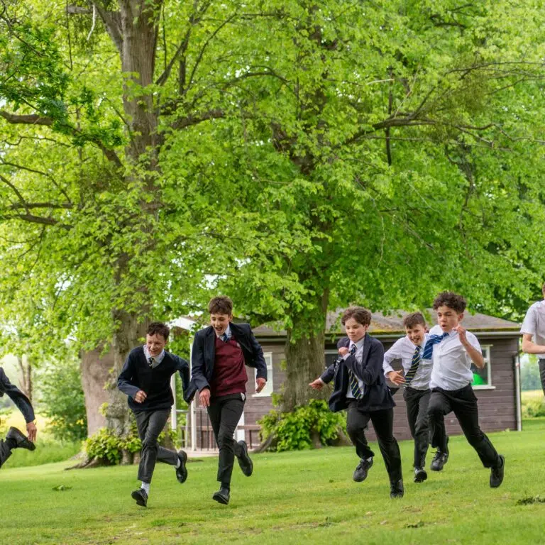 Group of school students in uniform running across a grassy yard under leafy trees.