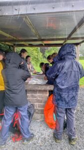 People in rain jackets gather around a stone table under a metal shelter, studying a large map or brochure.