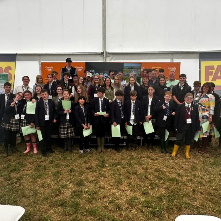 Group of students in school uniforms posing with adults inside a large tent at an outdoor event, holding green papers and badges.