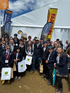 Group of students in school uniforms pose for a photo at an outdoor event, holding John Deere gift bags with a white tent and banners in the background.